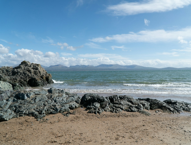 A sandy cove with rocks at Newborough Beach in Anglesey, looking across the sea at the mountains of Snowdonia.