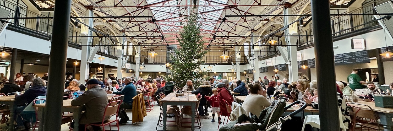 People seated at communal tables in front of a Christmas tree inside Glossop Market Hall.