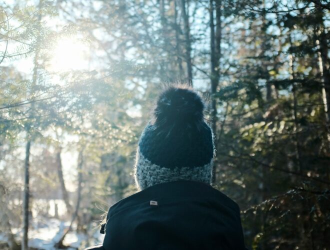 The back of a person walking in the snow in the woods with a bobble hat on.