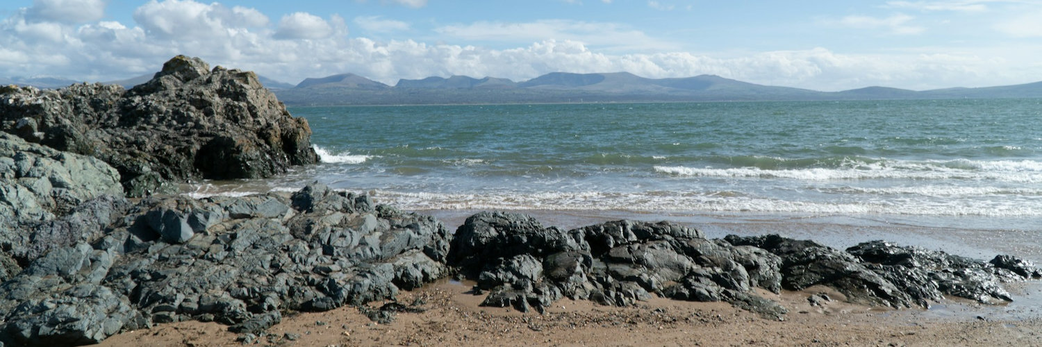 A sandy cove with rocks at Newborough Beach in Anglesey, looking across the sea at the mountains of Snowdonia.